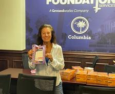 Woman smiling and holding a purple package in an office with boxes on a table behind her.