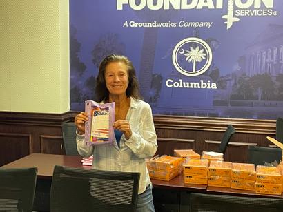 Woman smiling and holding a purple package in an office with boxes on a table behind her.
