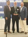 Three men in formal attire standing together, one holding an award, in front of a Triad Business Journal backdrop.