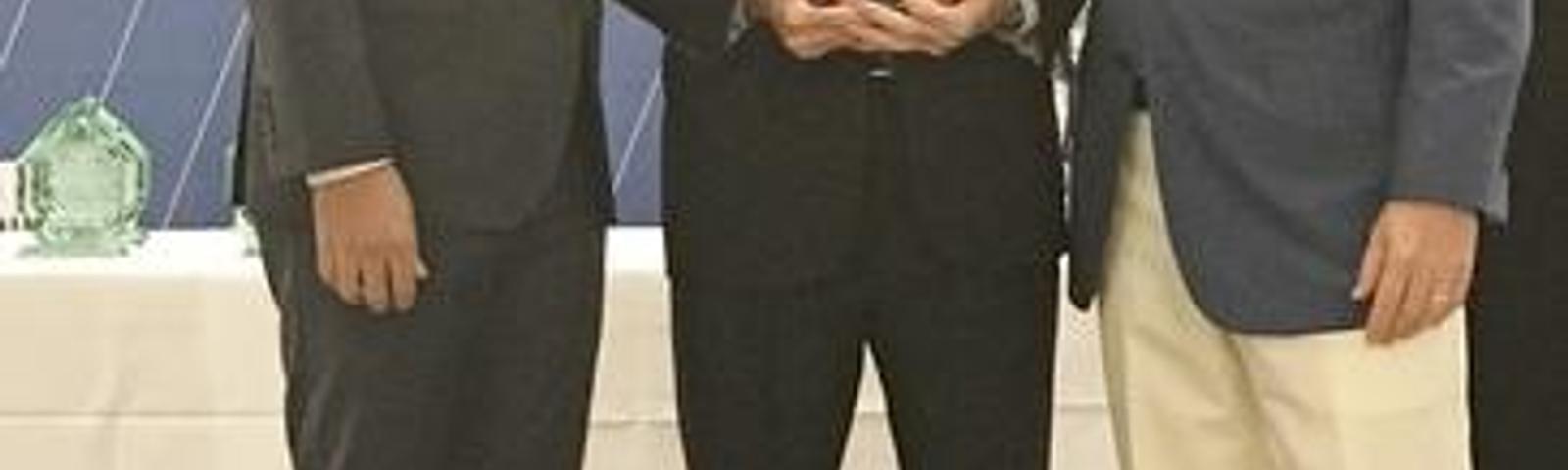 Three men in formal attire standing together, one holding an award, in front of a Triad Business Journal backdrop.
