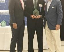 Three men in formal attire standing together, one holding an award, in front of a Triad Business Journal backdrop.