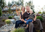 A smiling family of three sits outdoors in front of a house with a garden.