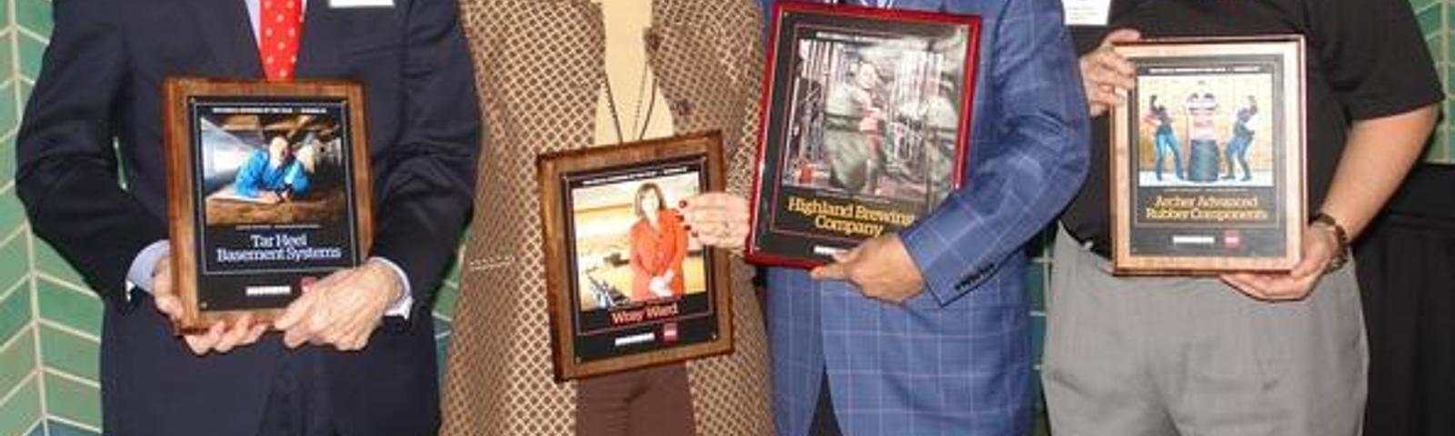 Four people standing and smiling, each holding framed awards in a decorated room.