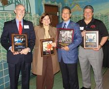 Four people standing and smiling, each holding framed awards in a decorated room.