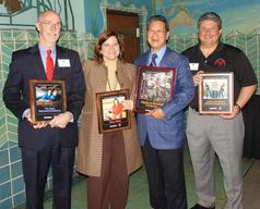 Four people standing and smiling, each holding framed awards in a decorated room.