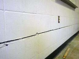 A cracked white wall with an electrical outlet and a piece of wood on the floor.