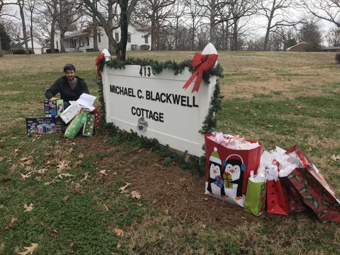 Man kneeling near a holiday-decorated sign with gifts, trees in the background.