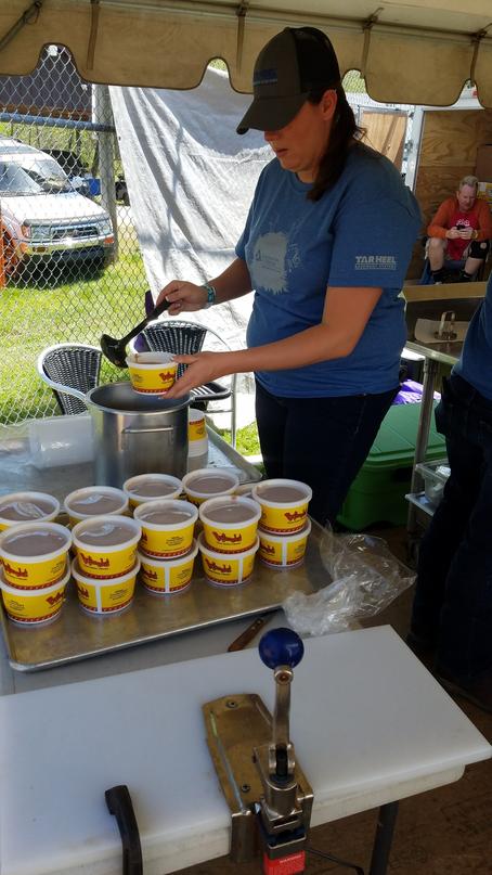 A person in a blue shirt serves food into yellow containers under a tent, supporting Communities in School.