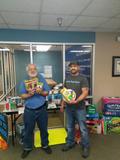 Two people holding school supplies in an office with more supplies stacked on a table and the floor.