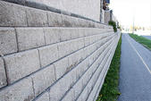 A long concrete retaining wall, serving as sturdy foundation walls, runs alongside a paved pathway with grass on the right.