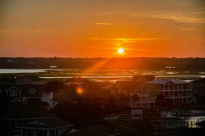 Sunset over a coastal town with houses in the foreground and orange sky reflected on water.