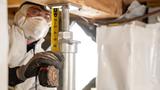 In protective gear, a person measures a support beam with a tape measure, essential for effective crawl space encapsulation.
