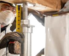 In protective gear, a person measures a support beam with a tape measure, essential for effective crawl space encapsulation.