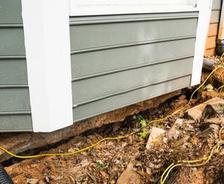 House foundation with visible cracks and erosion, surrounded by dirt and tangled yellow cables.