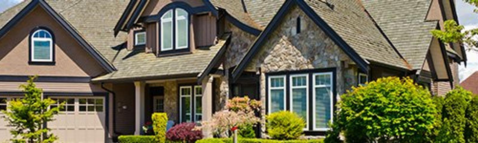 A large, modern house with stone accents, multiple peaked roofs, and a well-maintained garden in front.