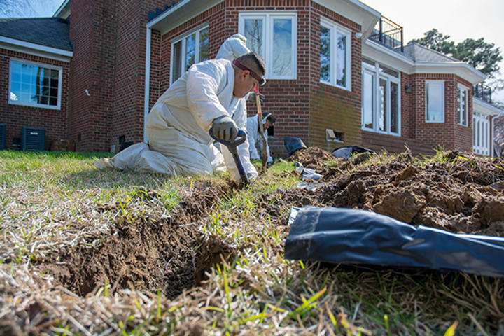 A person in protective clothing digs a trench in a grassy yard near a brick house.
