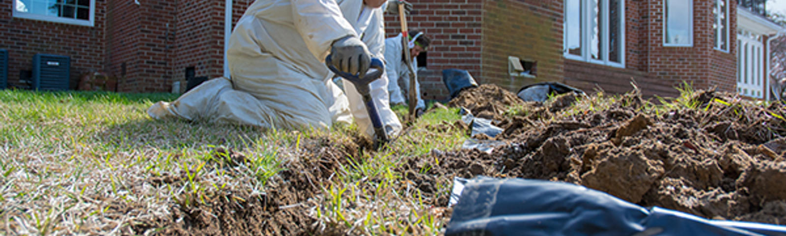A person in protective clothing digs a trench in a grassy yard near a brick house.