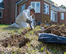 A person in protective clothing digs a trench in a grassy yard near a brick house.