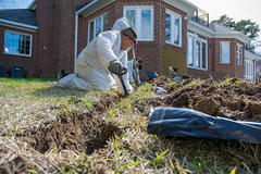 A person in protective clothing digs a trench in a grassy yard near a brick house.