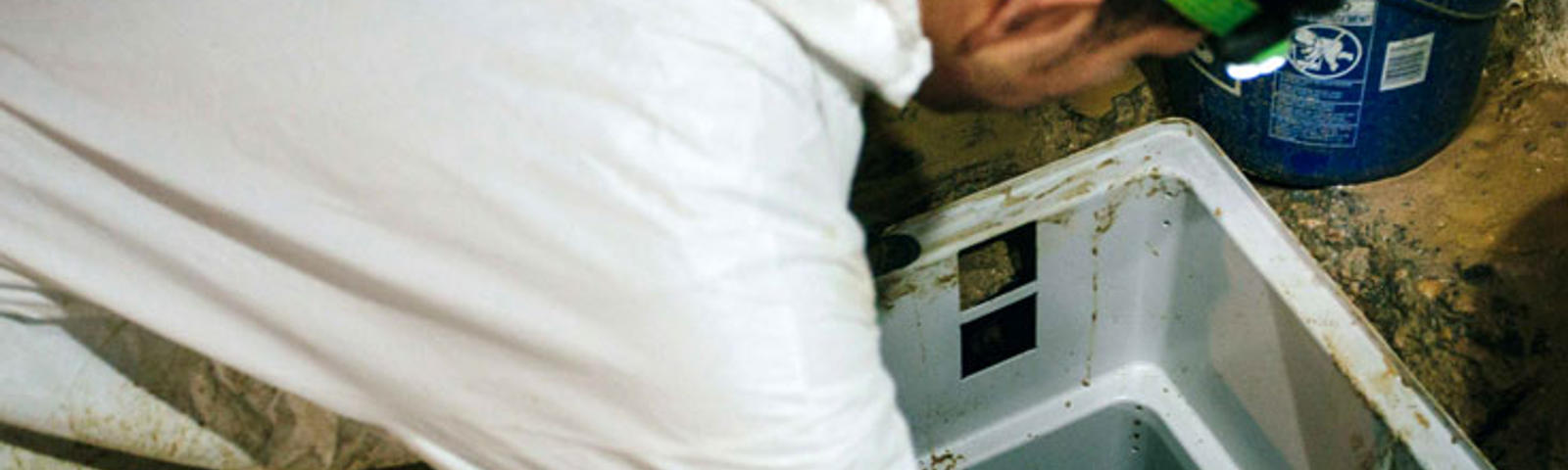 Person in white coveralls installing a sump pump in a basement with a blue bucket nearby.
