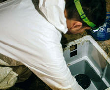 Person in white coveralls installing a sump pump in a basement with a blue bucket nearby.