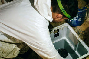 Person in white coveralls installing a sump pump in a basement with a blue bucket nearby.