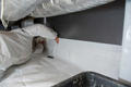 A person in a protective suit carefully works on installing insulation and a vapor barrier in the confined crawl space, with ducts looming overhead.