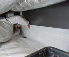 A person in a protective suit carefully works on installing insulation and a vapor barrier in the confined crawl space, with ducts looming overhead.