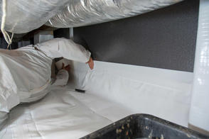 A person in a protective suit carefully works on installing insulation and a vapor barrier in the confined crawl space, with ducts looming overhead.