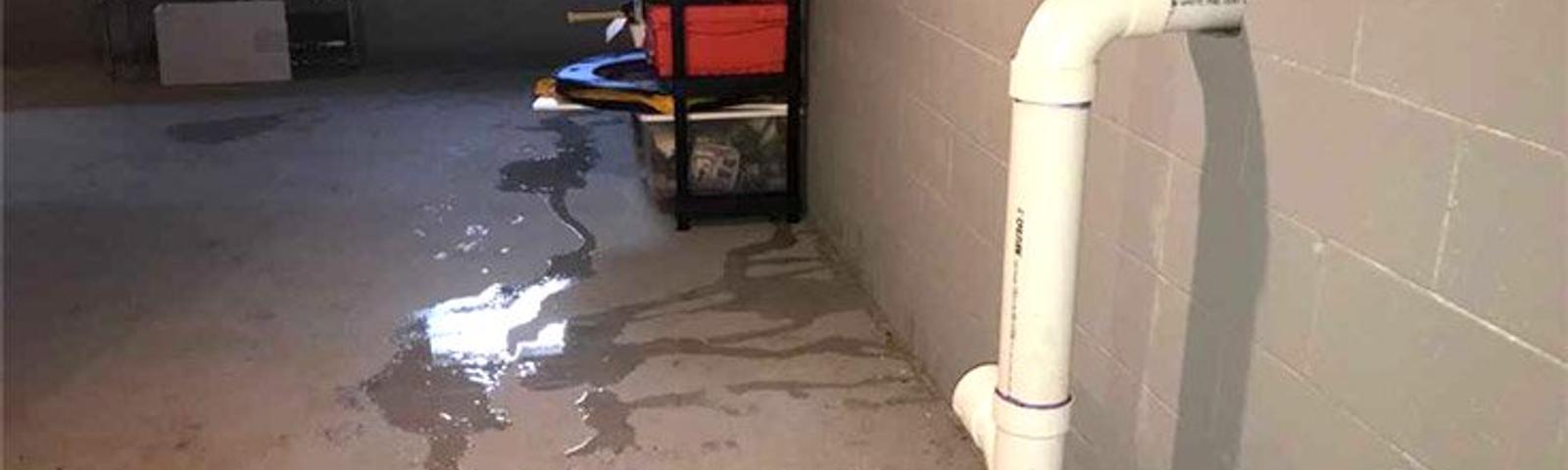 A basement with water leakage, showing puddles on the floor and a white pipe exiting the wall.