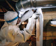 Person in protective suit and headlamp inspecting ductwork in a dimly lit space.