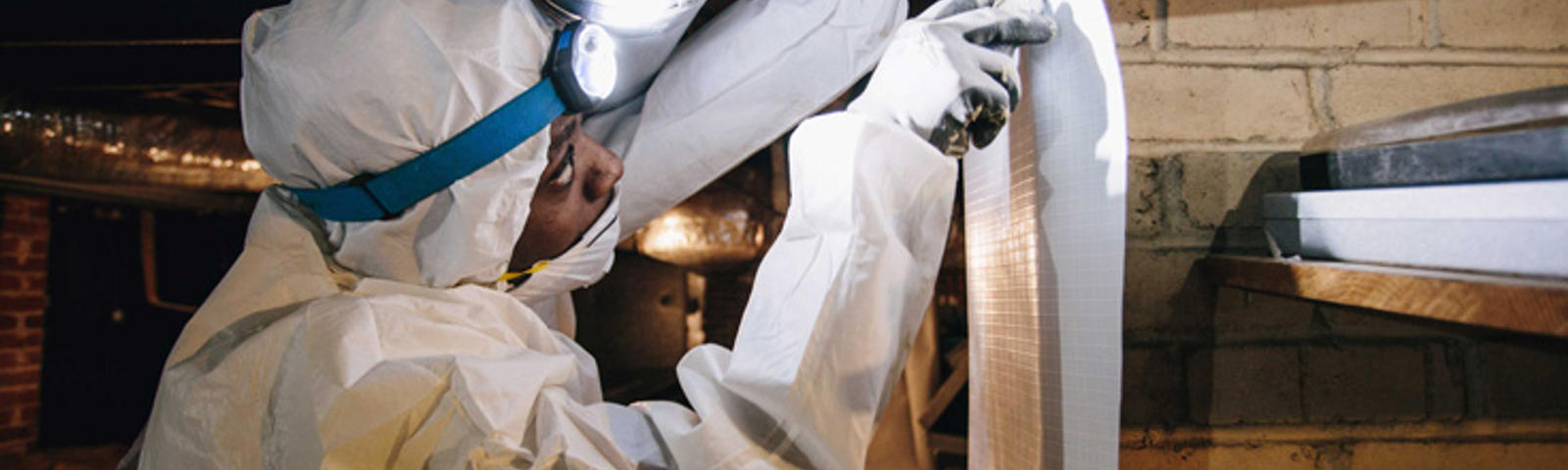 Person in protective suit and headlamp inspecting ductwork in a dimly lit space.
