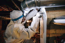 Person in protective suit and headlamp inspecting ductwork in a dimly lit space.