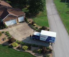 Aerial view of a moving truck parked in the driveway of a suburban house next to a quiet street.