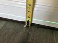 A hand measuring floor trim height with a tape measure against a beige wall, next to a green laser line on the floor.