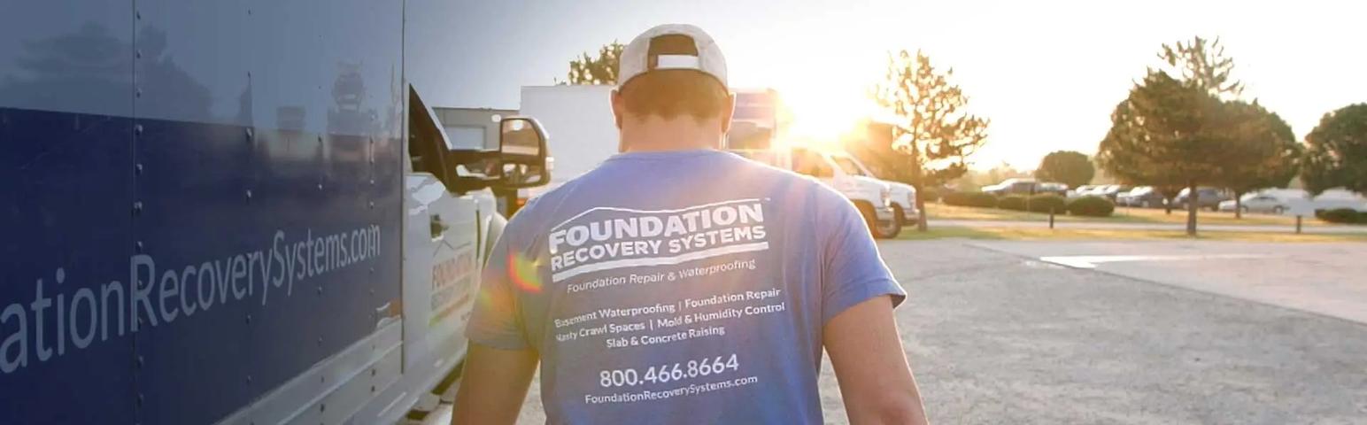Person in a blue "Foundation Recovery Systems" shirt walking towards vehicles in a sunny parking lot.