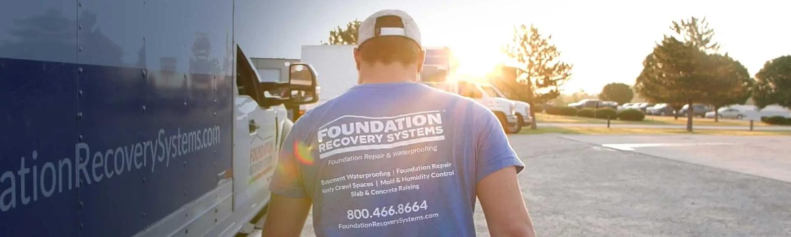 Person in a blue "Foundation Recovery Systems" shirt walking towards vehicles in a sunny parking lot.