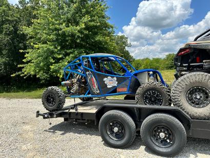 Blue off-road buggy on a trailer with a forested background under a partly cloudy sky.