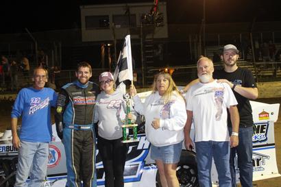 Group of six people celebrating by a race car, one holding a trophy, at an outdoor event at night.