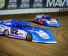 Two blue race cars on a dirt track, with number 22 leading another car. Advertising banners are in the background.