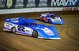 Two blue race cars on a dirt track, with number 22 leading another car. Advertising banners are in the background.