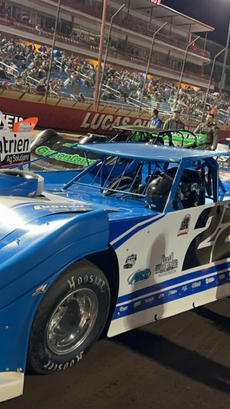 Race cars lined up on a track with drivers and crew members; spectators fill the stands in the background.