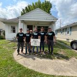 Five people in matching shirts hold a "Habitat for Humanity" sign in front of a house.