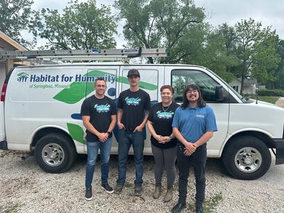 Four people stand in front of a Habitat for Humanity van, posing outdoors on a gravel surface.