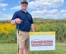 Smiling man holding a golf club stands next to a "Foundation Recovery Systems" sponsor sign in a field.