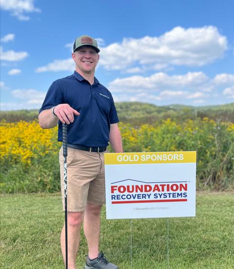 Smiling man holding a golf club stands next to a "Foundation Recovery Systems" sponsor sign in a field.