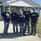 Five people wearing matching T-shirts stand smiling in front of a house on a sunny day.