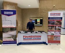 Man standing at a Foundation Recovery Systems display booth with banners and a table with brochures.