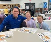 A smiling man and two young girls sit at a table with event programs in a crowded indoor setting.