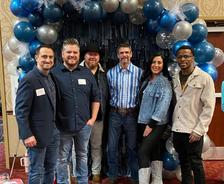 Six people stand smiling in front of a balloon arch at an indoor event.